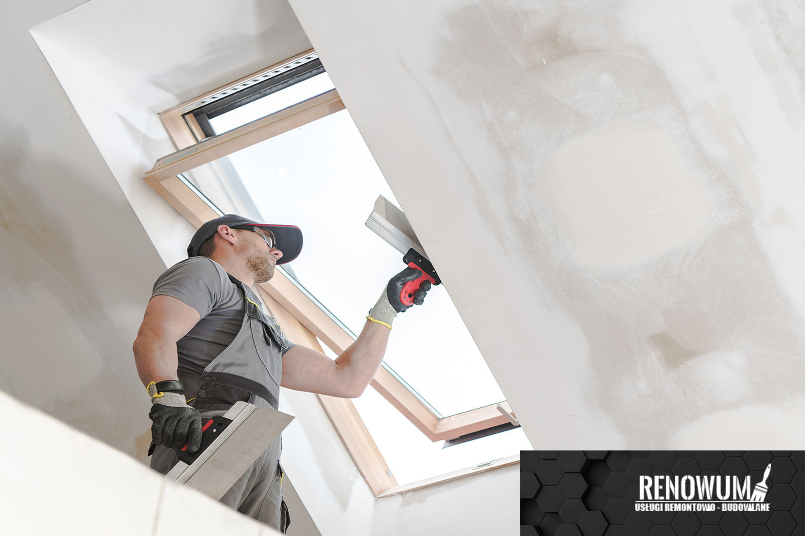 Pracownik w szarym kombinezonie i czapce wyrównuje szpachlą ścianę pod skosem przy oknie dachowym, widoczne ślady szpachlowania.