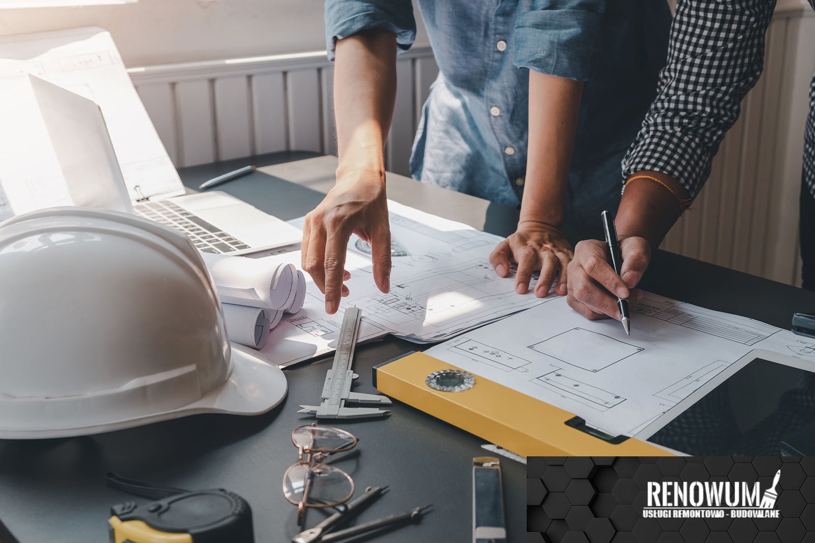 Dwóch architektów analizuje plan budynku na stole z kaskiem, laptopem, poziomica, suwmiarką, okularami i zwiniętymi rysunkami.