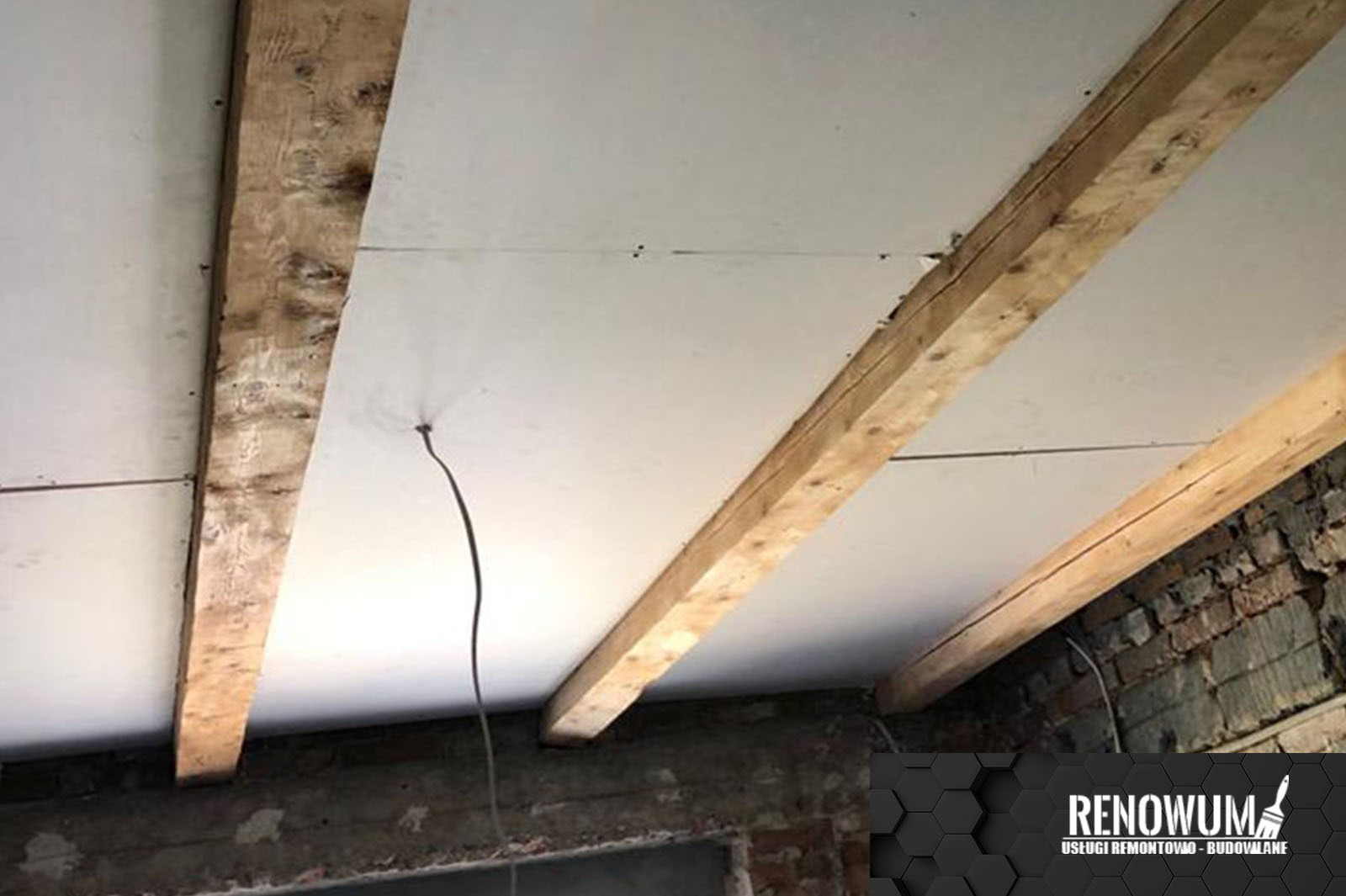 Płyty gipsowo-kartonowe zamontowane na drewnianych belkach stropowych, widoczne ślady po szpachlowaniu i wystający kabel elektryczny, fragment muru z cegły w tle.