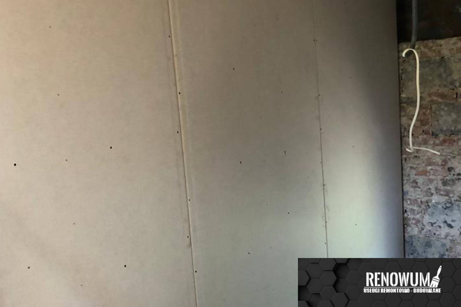 Wnętrze w trakcie prac: ściana wykończona płytami gipsowo-kartonowymi, widoczne wkręty, obok fragment starej ceglanej ściany z wystającym przewodem elektrycznym i logo firmy w rogu.