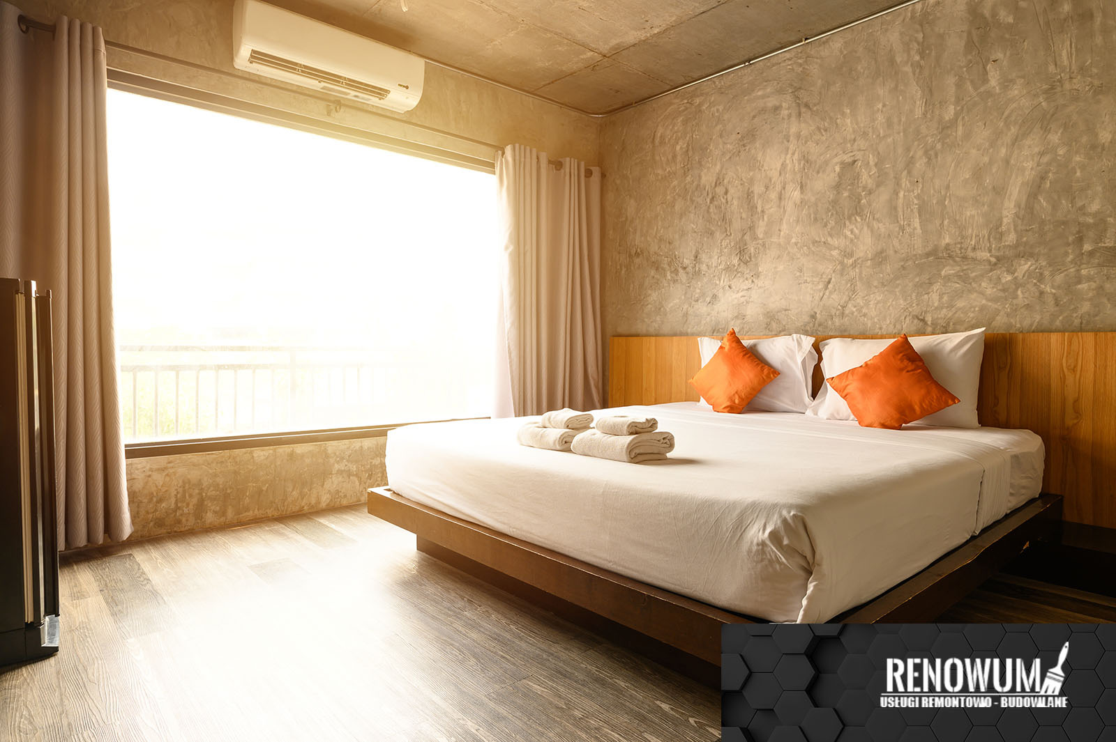 Nowoczesna sypialnia z surowym betonem na ścianie, jasnym oknem i minimalistycznym łóżkiem z pomarańczowymi poduszkami, widoczny klimatyzator i logo firmy w rogu kadru.
