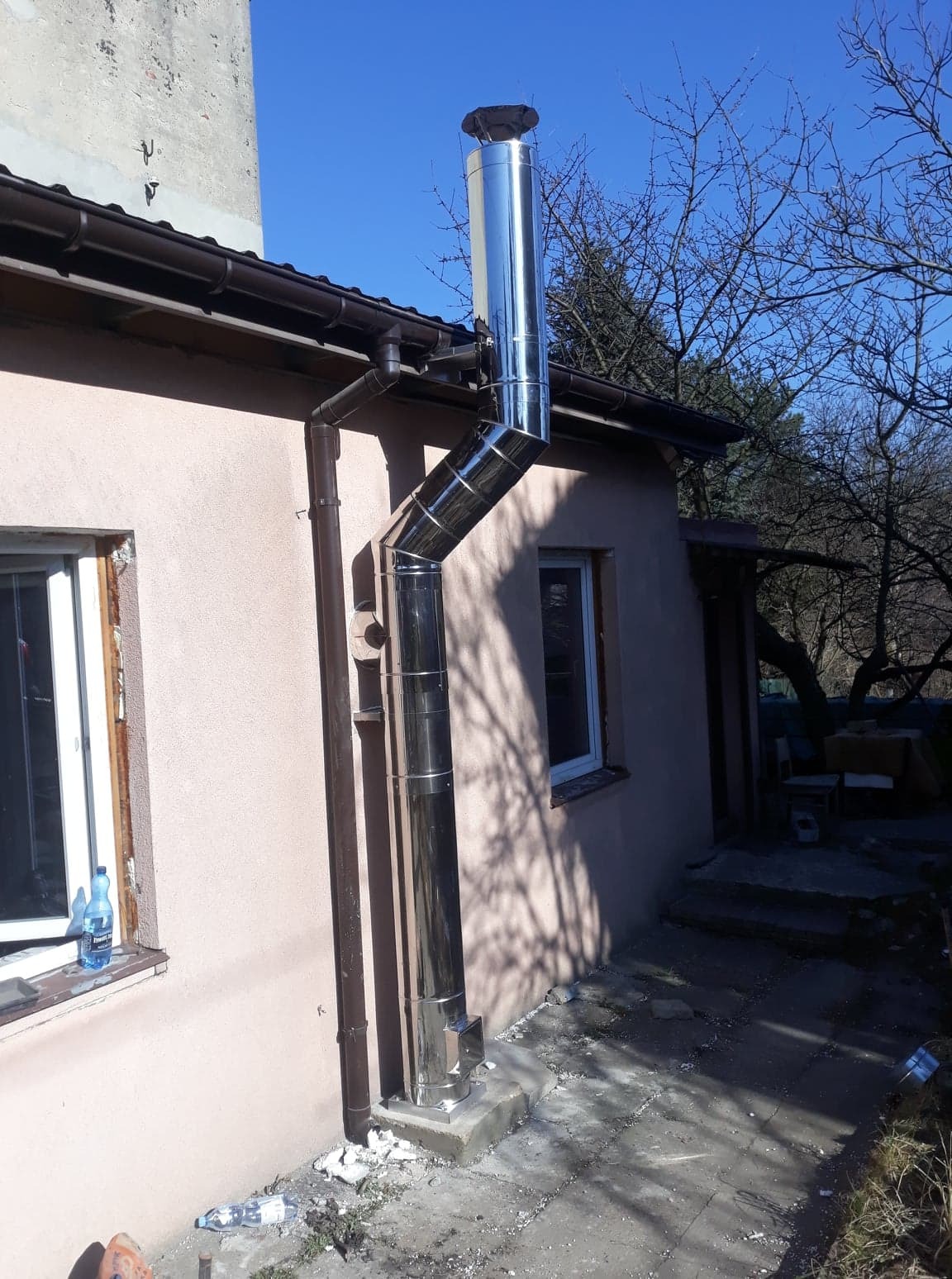 Zewnętrzny, błyszczący, stalowy system odprowadzania spalin z komina zamontowany na ścianie budynku, widoczne złącza segmentowe i daszek kominowy.