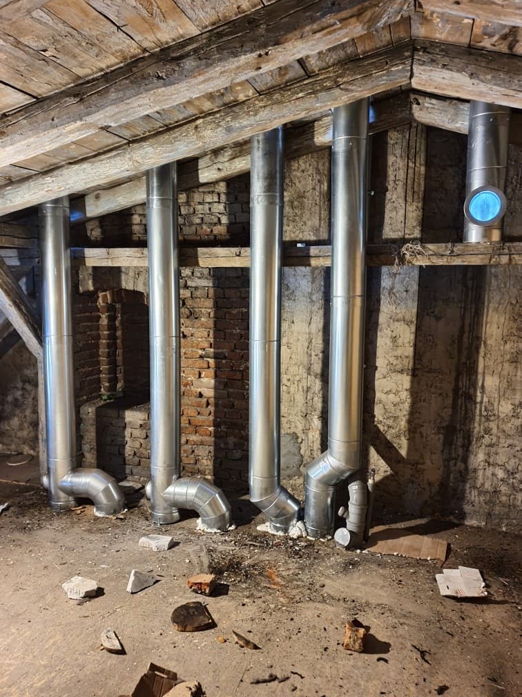 Montaż systemu odprowadzania spalin z pieca, widoczne rury spalinowe w surowym pomieszczeniu poddasza z drewnianą konstrukcją i fragmentami cegieł.