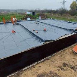 Dawid Kaleta - Prace ziemne przy budowie fundamentów: widok na wykop z izolacją, pracownik w czerwonej kurtce używa zagęszczarki, w tle łąka i drzewa.