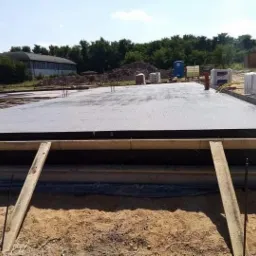 Zalany betonowy fundament z drewnianym szalunkiem i wystającymi prętami zbrojeniowymi, widok z bliska na piasek i deskę podpierającą szalunek.