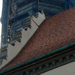 Dawid Kaleta - Fragment budynku z czerwoną dachówką, widoczne elementy dekoracyjne fasady i rusztowania z niebieską siatką w tle.