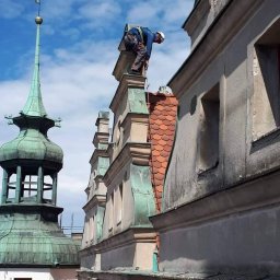 Dawid Kaleta - Pracownik w kasku i uprzęży zabezpieczającej na dachu zabytkowego budynku z wieżyczką, podczas prac na wysokości.