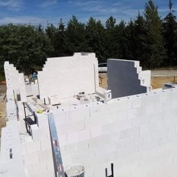 Dawid Kaleta - Budowa domu z białych bloczków betonu komórkowego, widoczne ściany w trakcie wznoszenia, poziomica oparta o ścianę, w tle las i błękitne niebo.