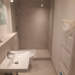 Dawid Kaleta - Nowoczesna łazienka w trakcie wykańczania, widoczna umywalka z baterią, toaleta, prysznic z szarą glazurą oraz grzejnik owinięty folią ochronną.