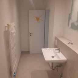 Dawid Kaleta - Nowa, jasna łazienka z umywalką wiszącą, grzejnikiem drabinkowym w folii ochronnej i niedokończonymi pracami malarskimi na ścianie w tle.