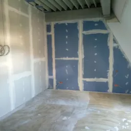 Wnętrze w trakcie prac wykończeniowych: ściany pokryte płytami kartonowo-gipsowymi, widoczne łączenia szpachlowane, wystające przewody elektryczne ze ścian. Podłoga częściowo zabezpieczona folią...