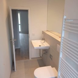 Dawid Kaleta - Nowoczesna łazienka z wiszącą toaletą, umywalką nablatową i dużym lustrem bez ramy, widok na otwarte drzwi i podłogę z szarych płytek.