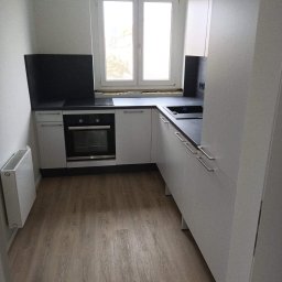 Dawid Kaleta - Jasna, minimalistyczna kuchnia w Brzegu z drewnianą podłogą, białymi szafkami i czarnym blatem, oświetlona naturalnym światłem z okna.