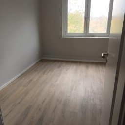 Dawid Kaleta - Pusty pokój z panelami podłogowymi w odcieniach beżu i szarości, szare ściany, białe listwy przypodłogowe i okno z widokiem na drzewa bez liści za szybą, uchylone drzwi po prawej stronie.