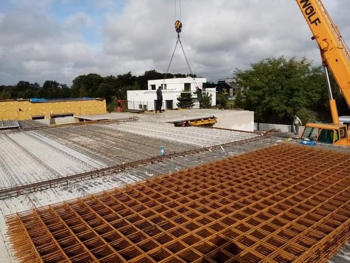 Budowa domu: widok z góry na zbrojenie stropu, z dźwigiem WOLF podnoszącym elementy konstrukcyjne na tle nowoczesnego budynku i zieleni.