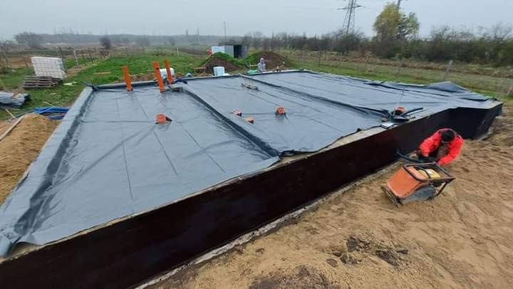 Prace ziemne przy budowie fundamentów: widok na wykop z izolacją, pracownik w czerwonej kurtce używa zagęszczarki, w tle łąka i drzewa.