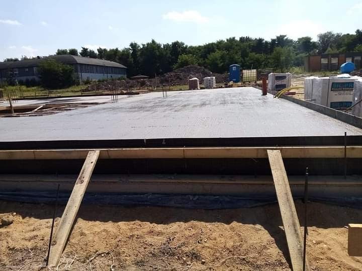 Zalany betonowy fundament z drewnianym szalunkiem i wystającymi prętami zbrojeniowymi, widok z bliska na piasek i deskę podpierającą szalunek.