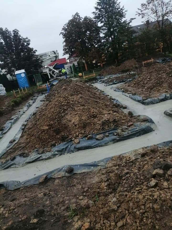 Wylane fundamenty pod budowę domu, widoczne szalunki i pryzmy ziemi, w tle betoniarka z wysięgnikiem i przenośna toaleta.