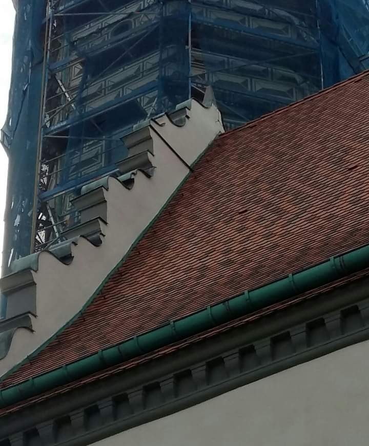 Fragment budynku z czerwoną dachówką, widoczne elementy dekoracyjne fasady i rusztowania z niebieską siatką w tle.