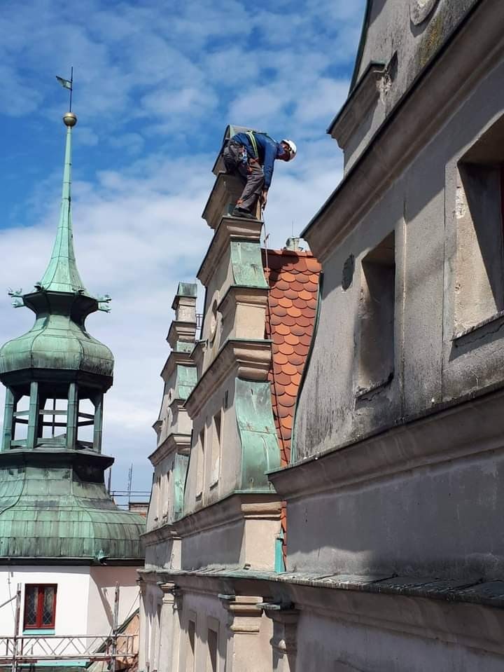Pracownik w kasku i uprzęży zabezpieczającej na dachu zabytkowego budynku z wieżyczką, podczas prac na wysokości.