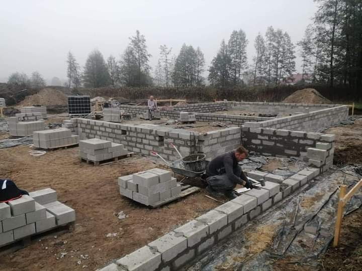 Budowa fundamentów domu z bloczków betonowych na zewnątrz, widoczny pracownik układający bloczki i palety z materiałem budowlanym w tle, pochmurna pogoda.