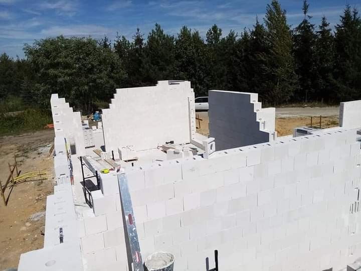 Budowa domu z białych bloczków betonu komórkowego, widoczne ściany w trakcie wznoszenia, poziomica oparta o ścianę, w tle las i błękitne niebo.