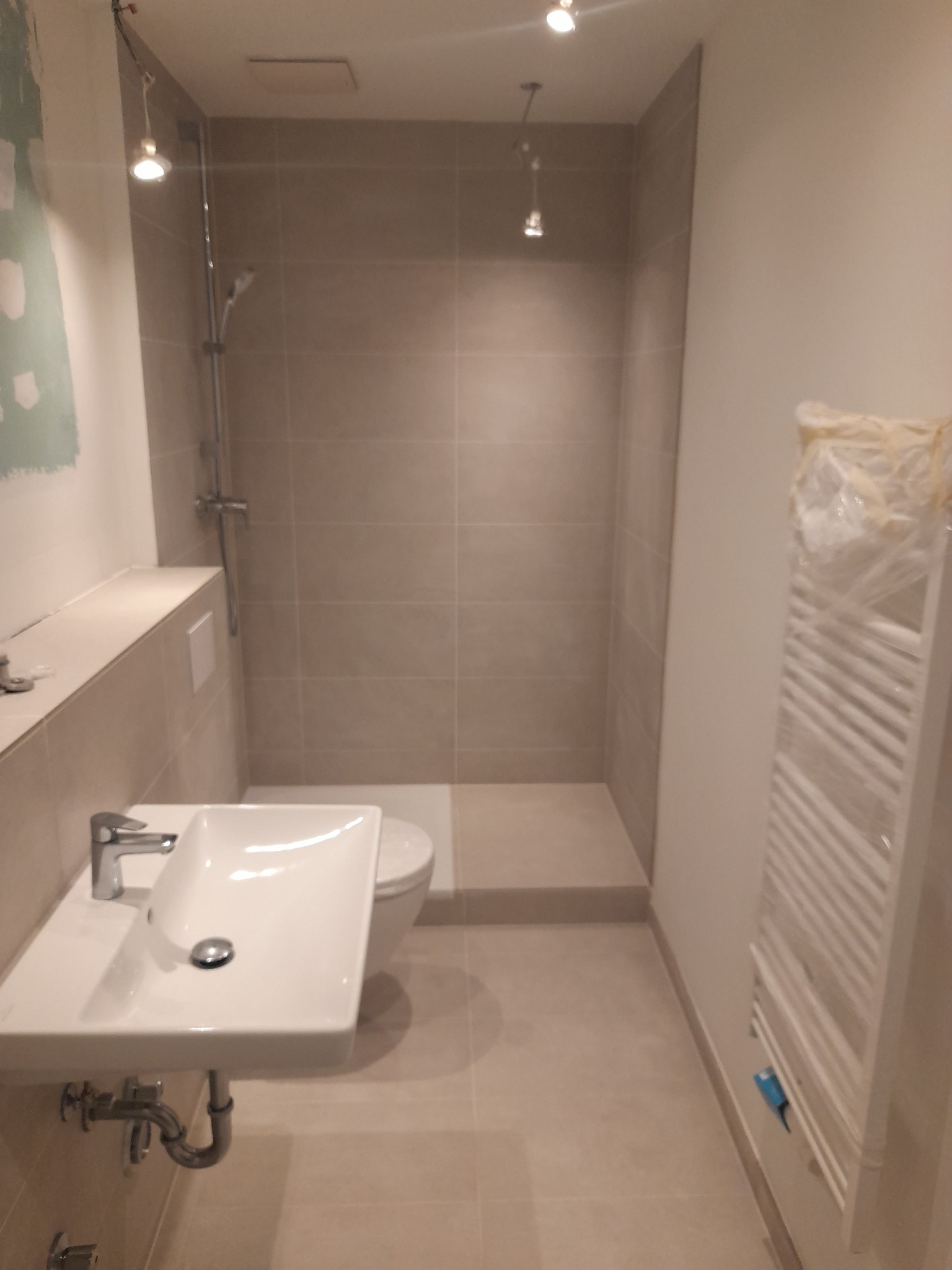 Nowoczesna łazienka w trakcie wykańczania, widoczna umywalka z baterią, toaleta, prysznic z szarą glazurą oraz grzejnik owinięty folią ochronną.