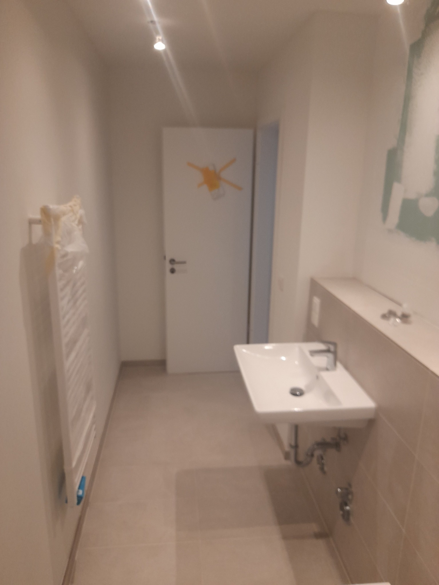 Nowa, jasna łazienka z umywalką wiszącą, grzejnikiem drabinkowym w folii ochronnej i niedokończonymi pracami malarskimi na ścianie w tle.