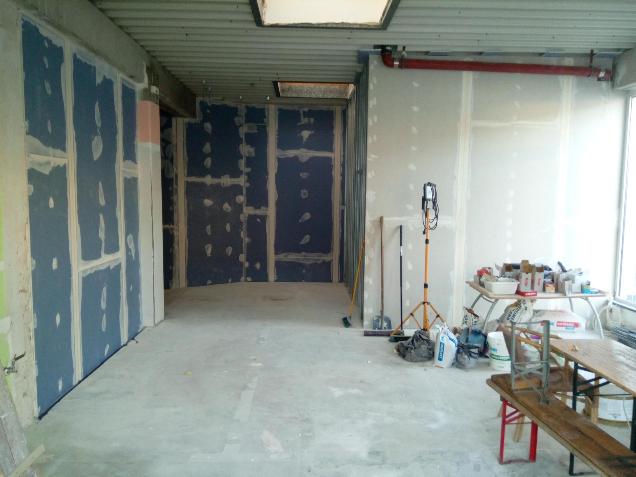 Wnętrze w trakcie montażu płyt kartonowo-gipsowych na stelażu, widoczne szpachlowane łączenia, narzędzia pracy i prowizoryczne stoły robocze z materiałami budowlanymi.