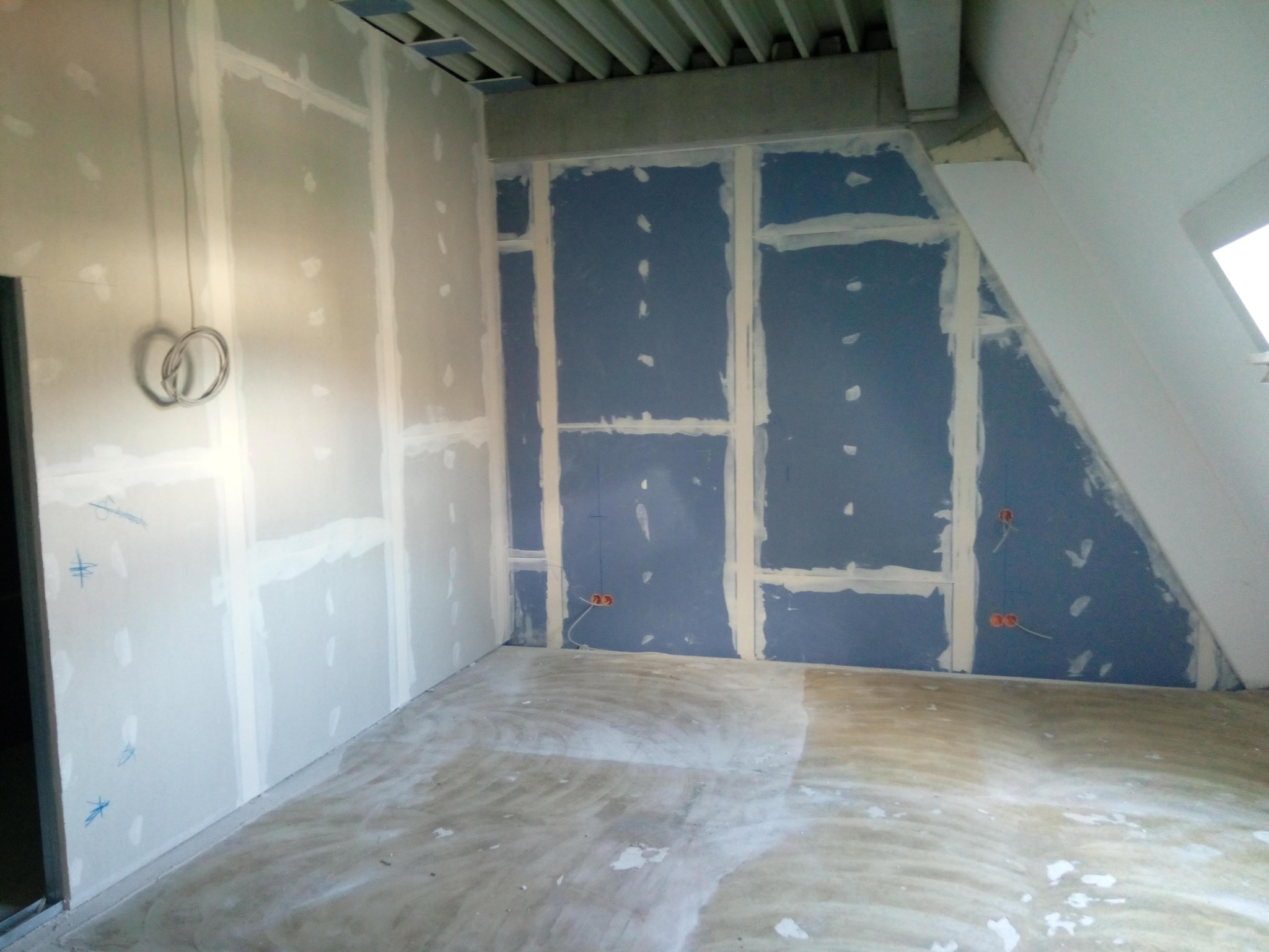 Wnętrze w trakcie prac wykończeniowych: ściany pokryte płytami kartonowo-gipsowymi, widoczne łączenia szpachlowane, wystające przewody elektryczne ze ścian. Podłoga częściowo zabezpieczona folią...