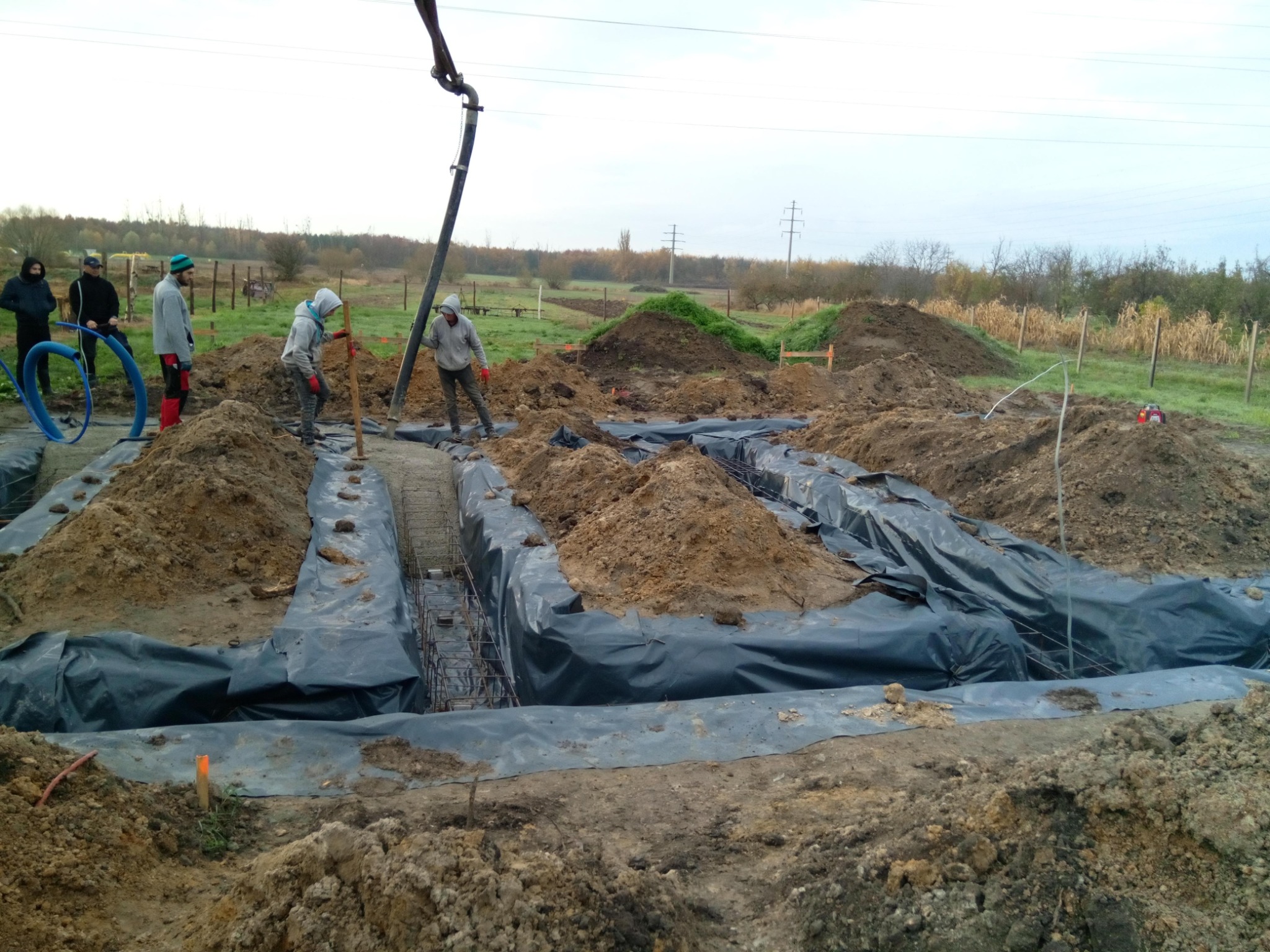 Przygotowanie wykopu pod fundamenty zbrojonego, z rozłożoną folią ochronną i ekipą budowlaną przy betonowaniu za pomocą pompy; w tle widoczne pryzmy ziemi i jesienny krajobraz.
