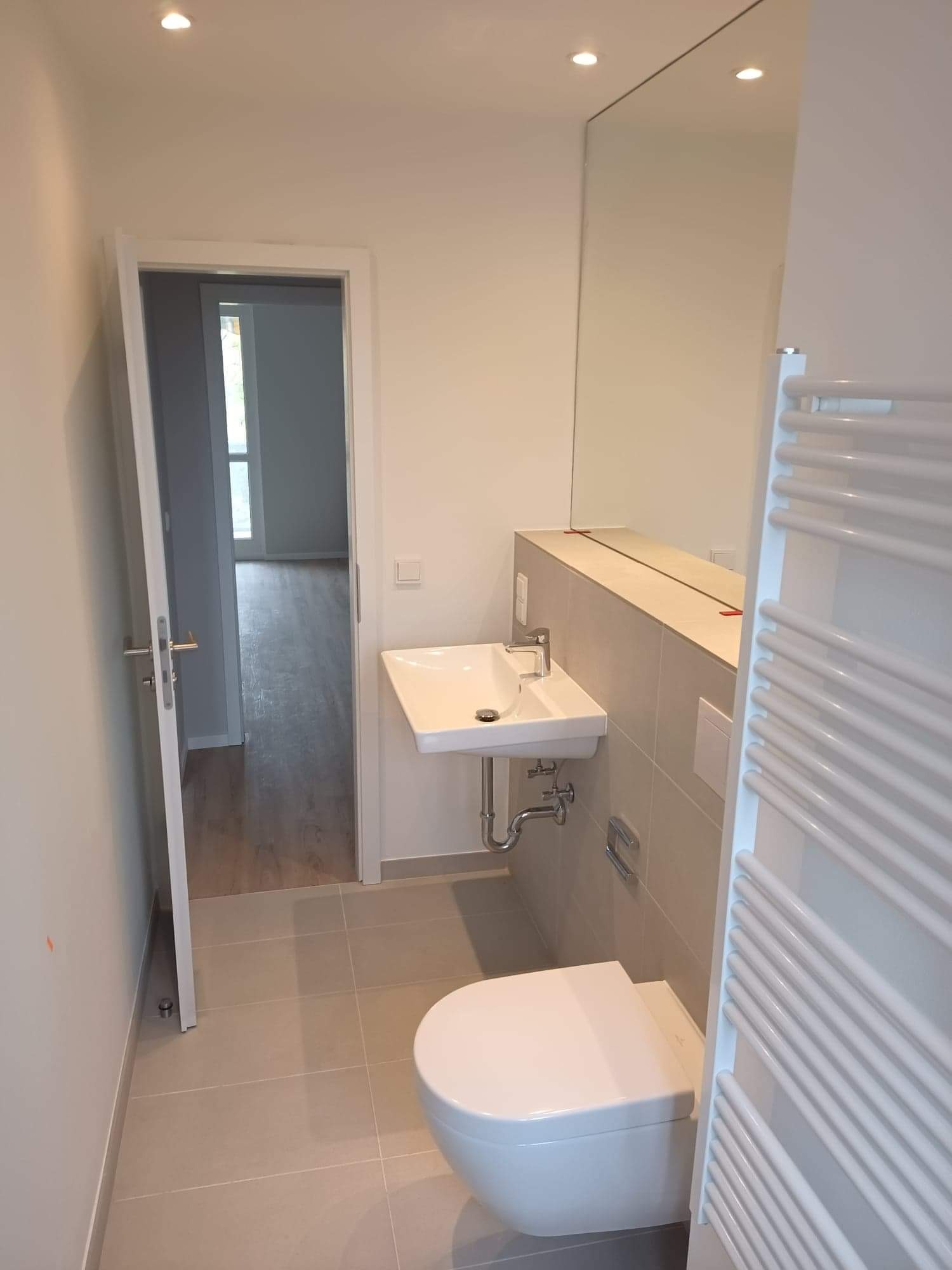 Nowoczesna łazienka z wiszącą toaletą, umywalką nablatową i dużym lustrem bez ramy, widok na otwarte drzwi i podłogę z szarych płytek.