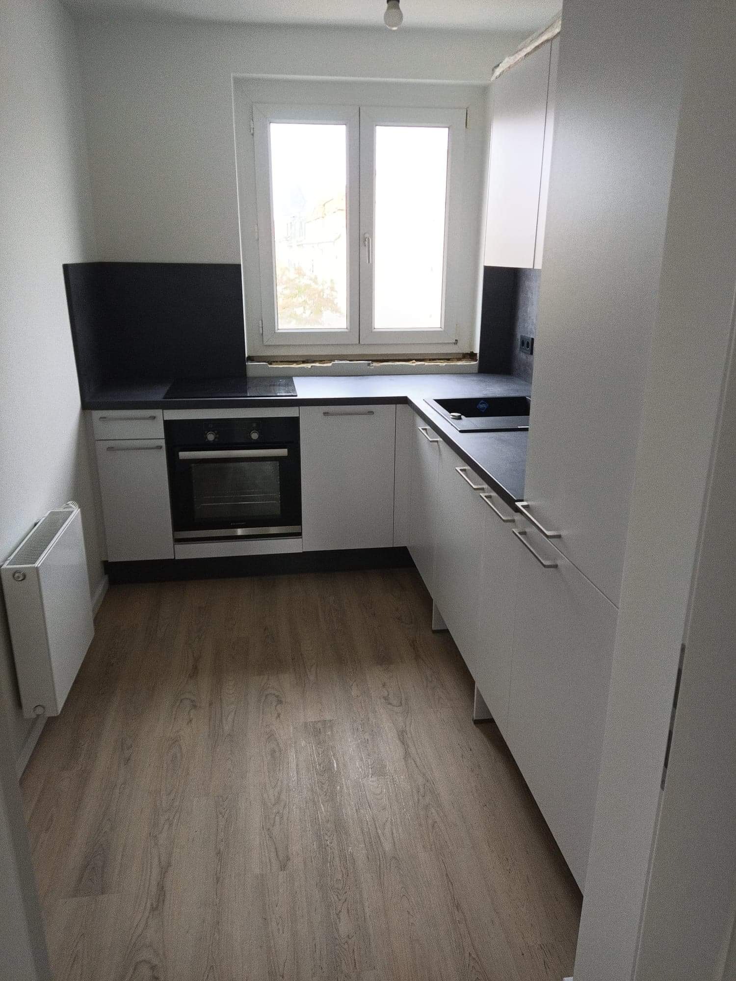 Jasna, minimalistyczna kuchnia w Brzegu z drewnianą podłogą, białymi szafkami i czarnym blatem, oświetlona naturalnym światłem z okna.