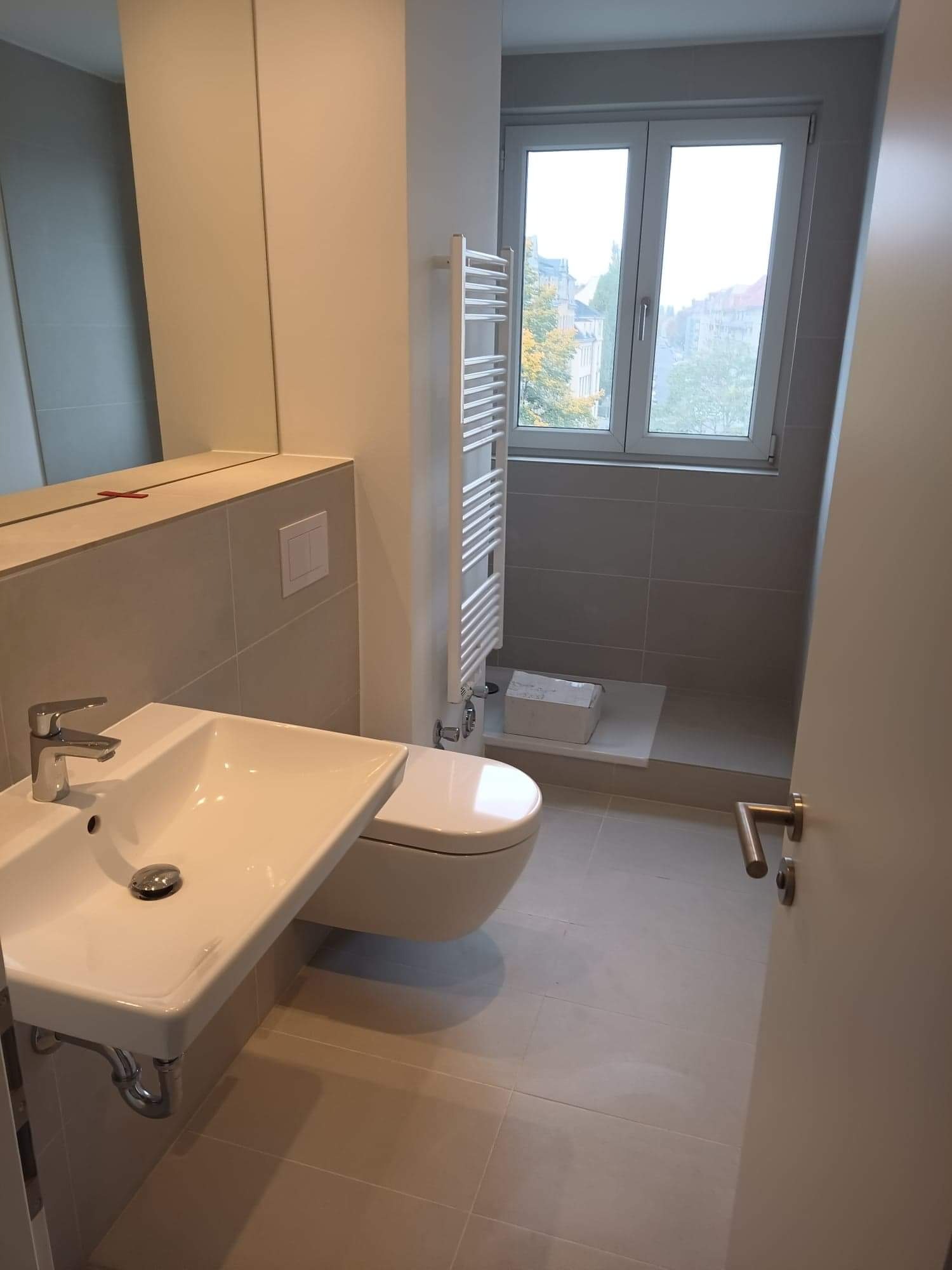 Nowoczesna łazienka w odcieniach szarości z wiszącą umywalką, toaletą i grzejnikiem drabinkowym; widok przez otwarte drzwi na zewnątrz z widocznymi drzewami.