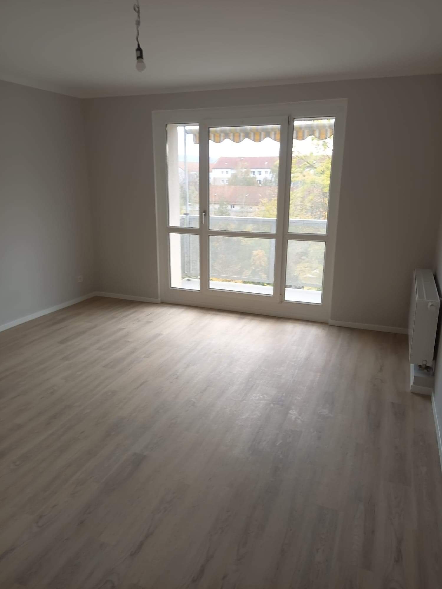 Pusty pokój po remoncie z panelami podłogowymi w odcieniach szarości, świeżo pomalowanymi ścianami na szaro, grzejnikiem i dużym oknem balkonowym z widokiem na budynki i drzewa.