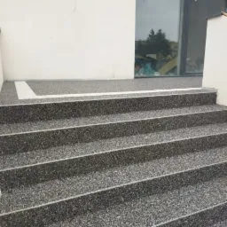 Schody zewnętrzne wykończone szarą, drobnoziarnistą posadzką żywiczną, z białym obramowaniem u góry, przylegające do białej ściany budynku z dużym oknem.