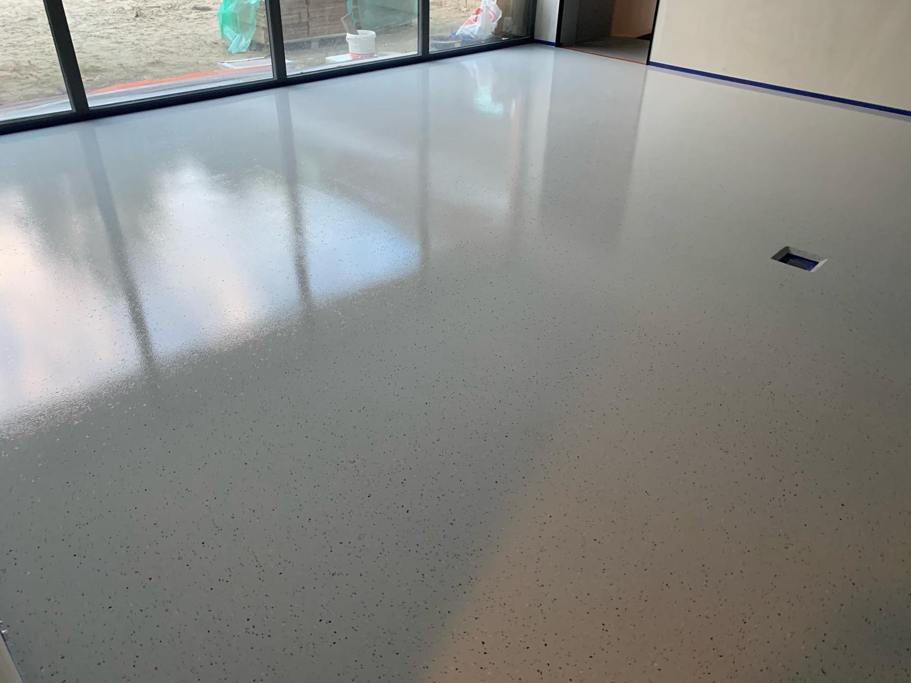 Jasnoszara, błyszcząca podłoga z drobnymi, ciemnymi wtrąceniami, widoczna kratka wentylacyjna w podłodze, taśma malarska przy ścianie, duże okno z widokiem na zewnątrz.