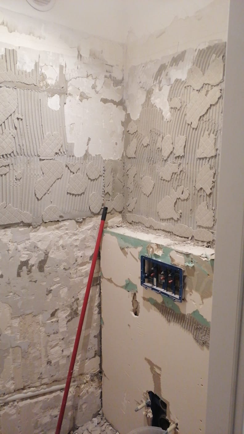 Przygotowanie ścian w łazience do położenia płytek, widoczne resztki starej okładziny, nałożony klej z charakterystycznym wzorem, czerwony kij oparty o ścianę, instalacja wodna w niebieskiej obudowie.