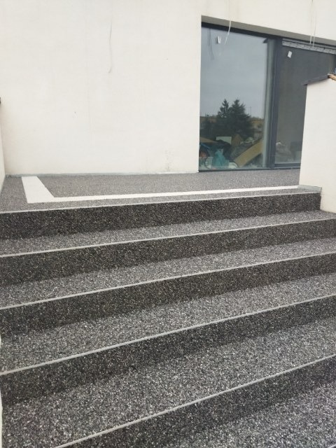 Schody zewnętrzne wykończone szarą, drobnoziarnistą posadzką żywiczną, z białym obramowaniem u góry, przylegające do białej ściany budynku z dużym oknem.
