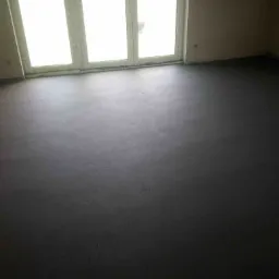 Świeżo wylana, ciemnoszara posadzka w pustym pomieszczeniu z dużym oknem balkonowym. Widoczne ślady po wyrównywaniu powierzchni.