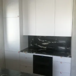 Nowoczesna zabudowa kuchenna z białymi szafkami, czarnym blatem z marmurkowym wzorem i miedzianymi uchwytami, z piekarnikiem w zabudowie.