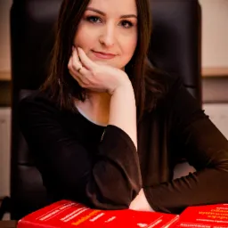 Portret kobiety w czarnej bluzce, opierającej brodę na dłoni z pierścionkiem, za nią czarne krzesło biurowe, na pierwszym planie dwa czerwone tomy komentarzy do kodeksu cywilnego.