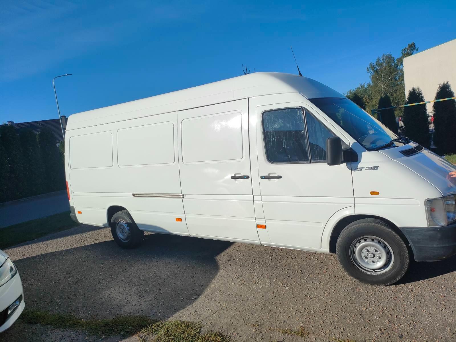 Biały samochód dostawczy Volkswagen LT 35 zaparkowany na żwirowym podłożu, zarys innego pojazdu widoczny z lewej strony.