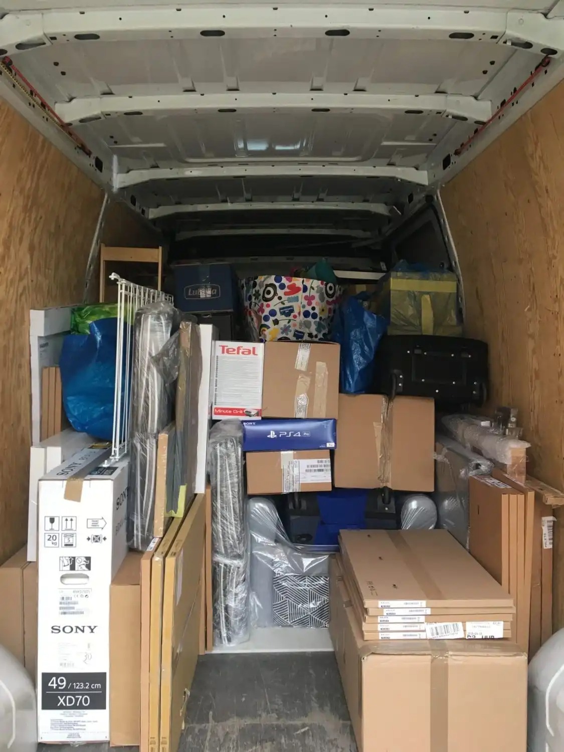 Wnętrze furgonetki wypełnionej kartonami i przedmiotami, w tym pudełka Sony TV, Tefal i PS4, całość zabezpieczona do transportu.