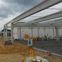 Budowa hali: widoczny szkielet konstrukcji z betonu, zbrojenie pod wylewkę, w tle pracownicy w kamizelkach odblaskowych, na pierwszym planie niwelator na statywie i hałda piachu.