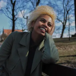 Portret uśmiechniętej kobiety z blond afro w zielonym płaszczu, opierającej się o barierkę na tle błękitnego nieba i drzew bez liści.