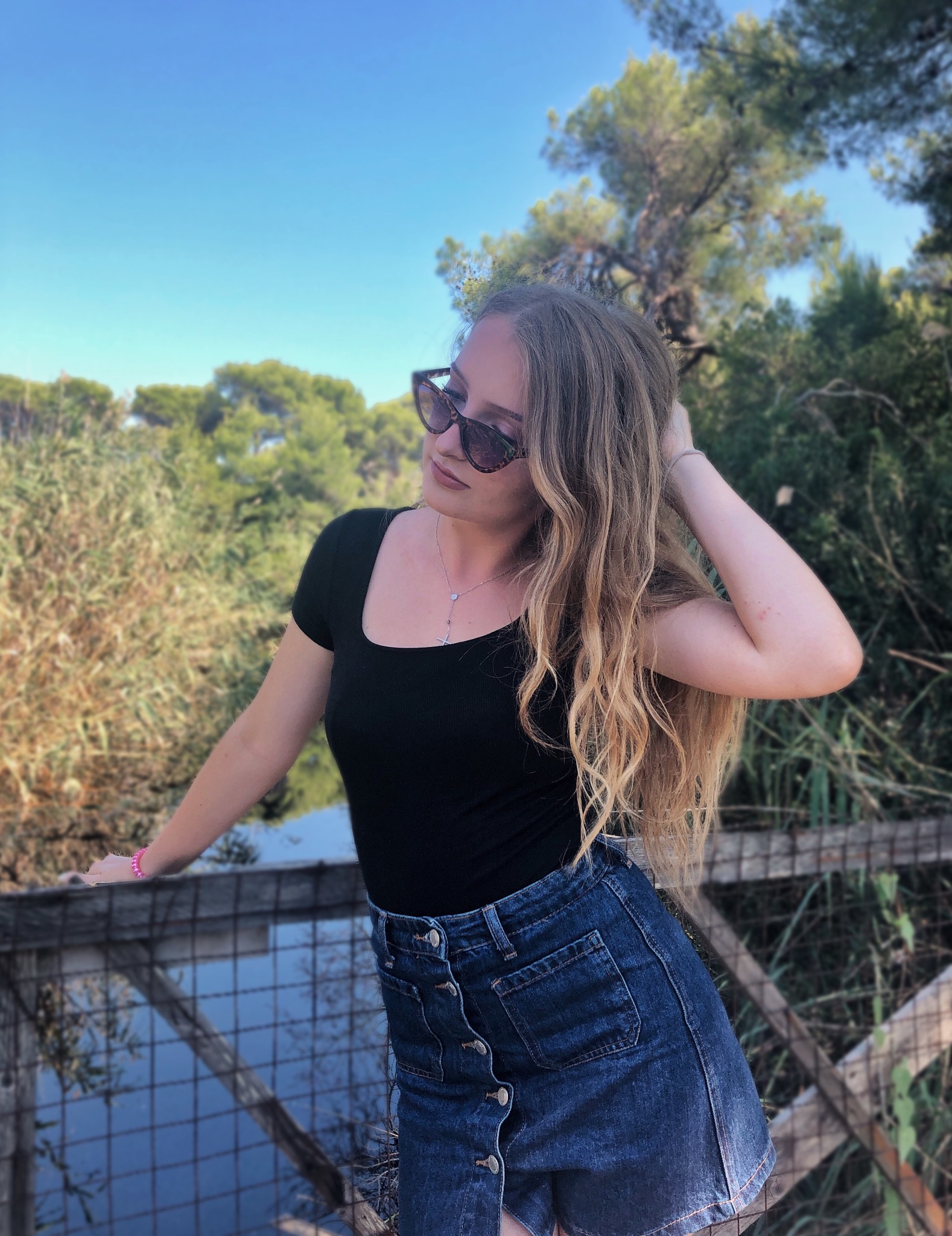 Kobieta w czarnej bluzce i dżinsowej spódnicy pozuje na tle zieleni i błękitnego nieba, opierając się o drewnianą barierkę.