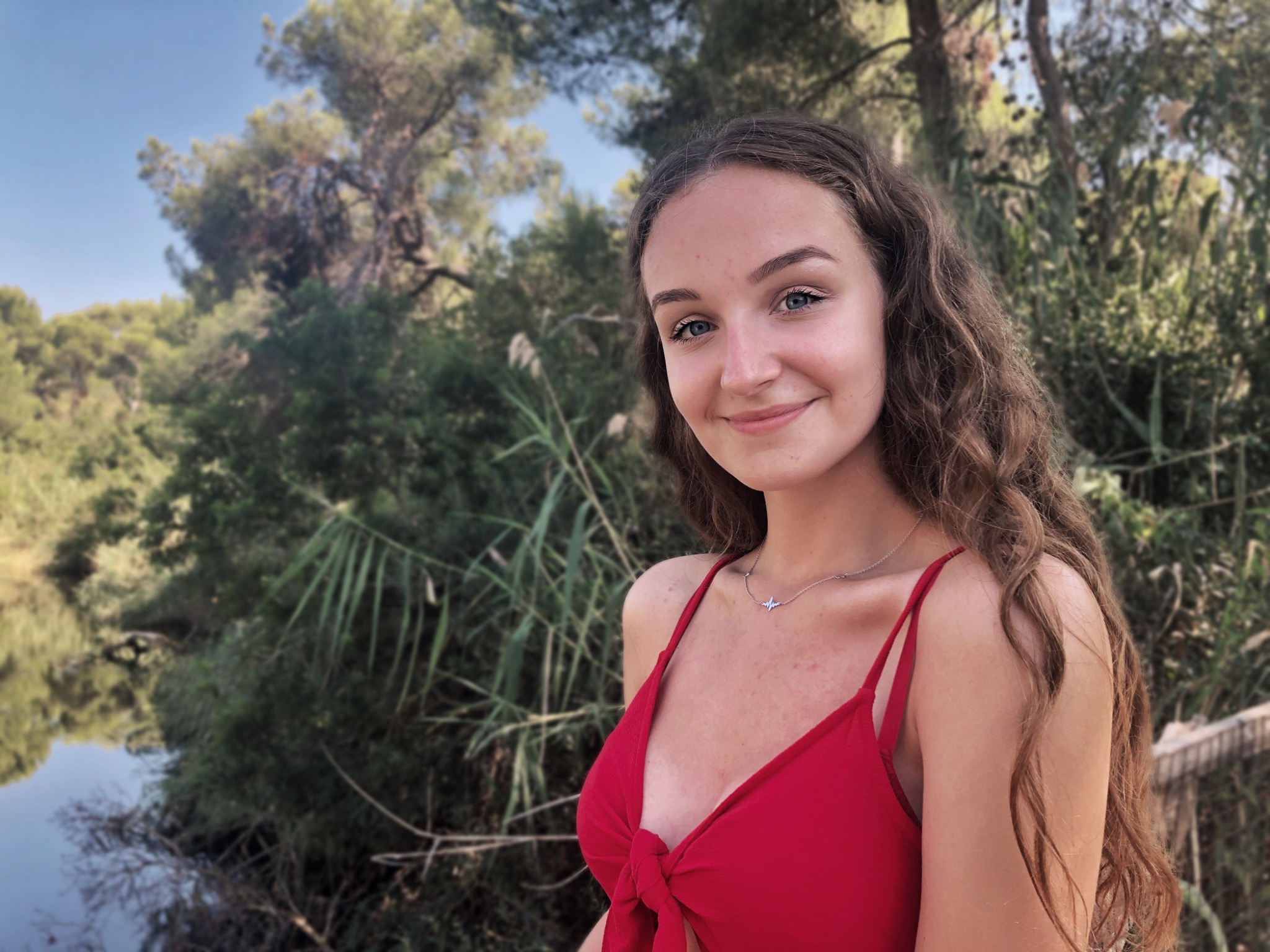 Portret uśmiechniętej, młodej kobiety w czerwonym stroju na tle zielonej roślinności i wody, z delikatnym naszyjnikiem w kształcie linii życia.