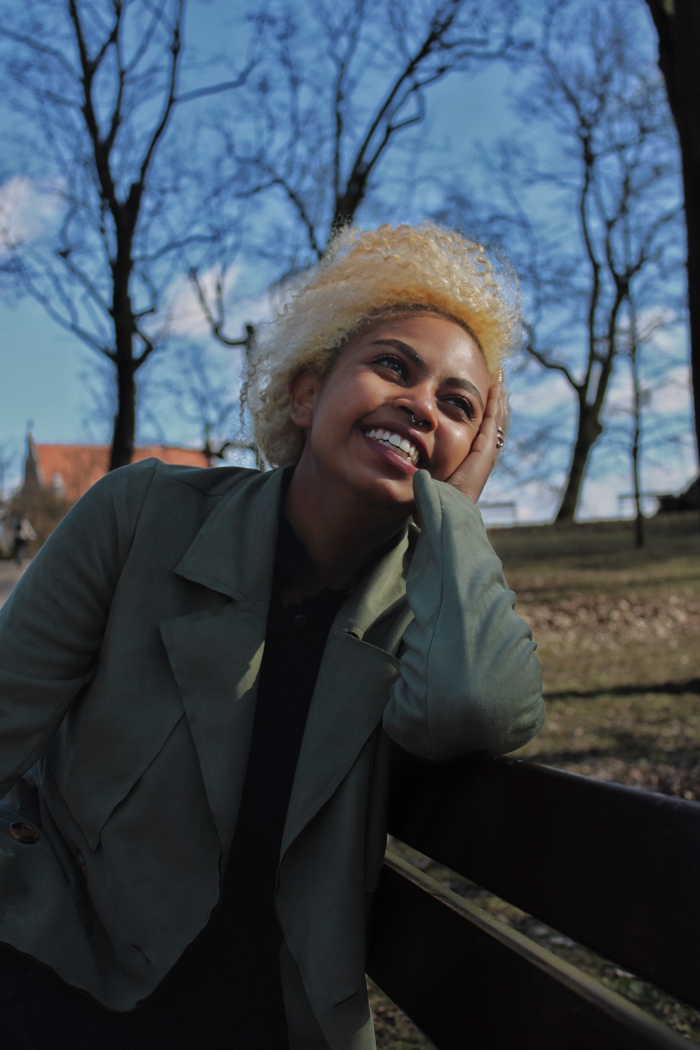 Portret uśmiechniętej kobiety z blond afro w zielonym płaszczu, opierającej się o barierkę na tle błękitnego nieba i drzew bez liści.