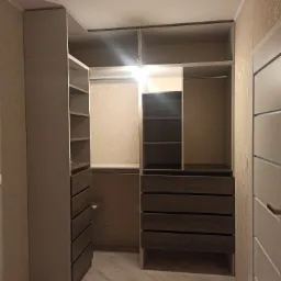 Wnętrze garderoby z szufladami i półkami w odcieniach szarości, oświetlone górnym światłem, z widoczną fakturą tapety na ścianie i fragmentem białych drzwi po prawej stronie.